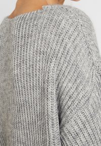 Ljusgrå stickad tröja med ribbat mönster, med en bred halsringning och en avslappnad passform, som visar ett intrikat mönster längs ärmarna.