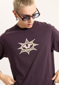 Purple cotton t-shirt featuring a central graphic with a stylized "V" in an oval, surrounded by dynamic green and black checkerboard accents.