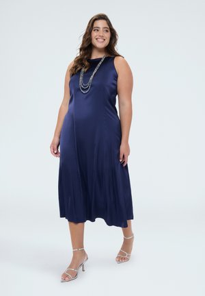 Lächelnde Frau, die ein ärmelloses, marineblaues Satin-Midikleid trägt, eine mehrlagige Halskette und silberne Sandalen mit hohen Absätzen, stehend vor einem schlichten Hintergrund.