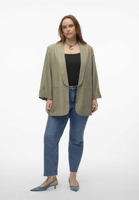 Blazer vert olive avec des manches retroussées, associé à un haut noir, un jean bleu taille haute et des escarpins pointus bleu clair. Détail de collier simple.