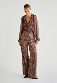 Musterhaftes, mehrfarbiges Outfit, bestehend aus einer lockeren Bluse mit Rüschen und hochtaillierten, weiten Hosen, beide in lila, goldenen und braunen Farbtönen.