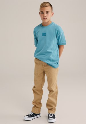 Camisa de algodón turquesa con un diseño gráfico azul, combinada con pantalones caqui claros y zapatillas negras y blancas, con un corte relajado.