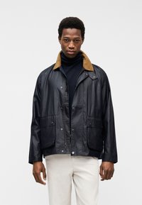 Νέος άνδρας που φοράει μαύρο μπουφάν με καφέ γιακά, μαύρο ζιβάγκο και ανοιχτόχρωμο παντελόνι, στέκεται μπροστά σε απλό λευκό φόντο.