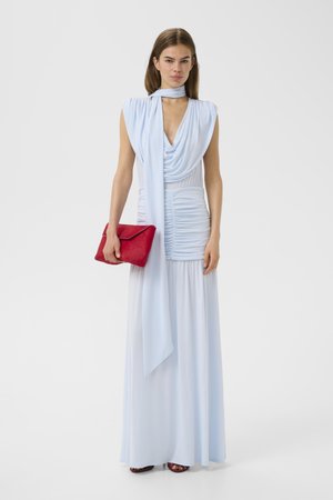 Kvinne iført en ermeløs, lyseblå maksi kjole med rysjedetaljer og et matchende skjerf, holder en rød clutch-veske, stående på hvit bakgrunn.