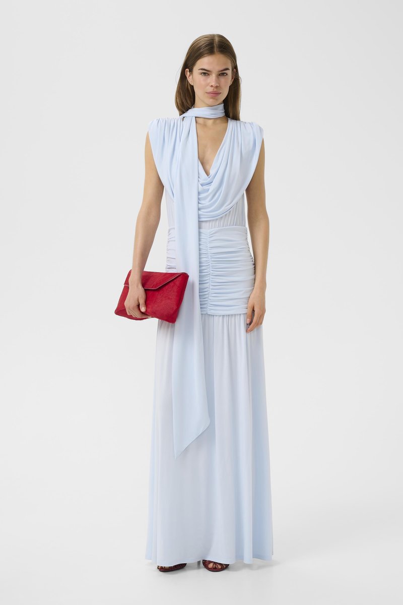 Γυναίκα που φοράει ένα αμάνικο, φουσκωτό maxi φόρεμα σε ανοιχτό γαλάζιο χρώμα με matching φουλάρι, κρατώντας μια κόκκινη τσάντα φάκελο, στέκεται σε λευκό φόντο.