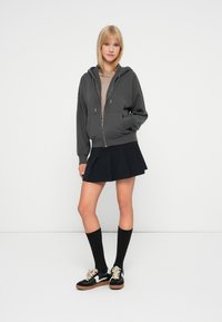 Siva zip-up dukserica s ležernim krojem, složena preko bež majice. Uparena s crnom plisiranom suknjom, čarapkama do koljena crne boje i crnim tenisicama.