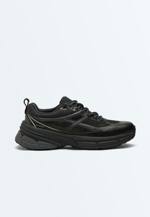 Black chunky sneaker with thick sole, mesh and synthetic upper, padded collar, lace-up front, and pull tab on heel, shown in side view.