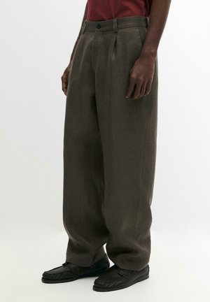 Pantaloni ampi color oliva scuro indossati da una persona con pelle scura, abbinati a scarpe nere e a una camicia rosso scuro infilata dentro.