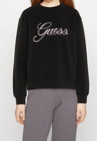 Svart sweatshirt med ribbad textur, utsmyckad med en glittrande "Guess"-logotyp i silver och svarta detaljer, rund hals och elastiska manschetter.