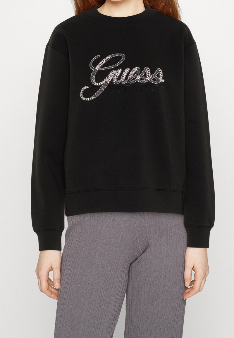 Svart sweatshirt med ribbad textur, utsmyckad med en glittrande "Guess"-logotyp i silver och svarta detaljer, rund hals och elastiska manschetter.