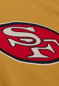 Gelbes Material zeigt ein auffälliges rotes und weißes ovales Logo mit schwarzen Umrissen, das die Buchstaben "SF" in einer fetten, stilisierten Schrift darstellt.