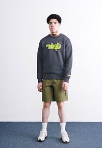 Γκρίζα φόρμα με νεον πράσινο λογότυπο "Nike", συνδυασμένη με ελιόχρωμες βερμούδες. Λευκές κάλτσες και αθλητικά παπούτσια ολοκληρώνουν το σύνολο. Απλός σχεδιασμός.