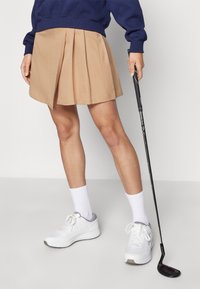 Beige pliszírozott golf szoknya, pamut keverékből, sima textúrával. Fehér tornacipővel párosítva, és egy golfütő mellett elhelyezve.