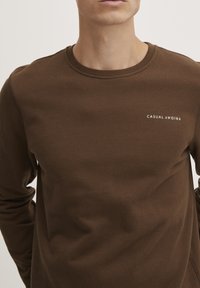 Brun langermet genser i et mykt stoff med rund hals. Har hvit tekst "CASUAL FRIDAY" på brystet, minimalistisk design.