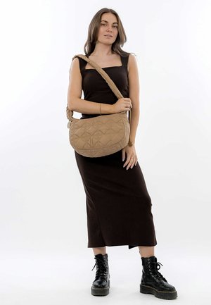 Gepolsterte beige Schultertasche mit geschwungener Form, geflochtenem Riemen und subtilm Rautenmuster. Das Modell trägt ein enges braunes Kleid und schwarze Stiefel.
