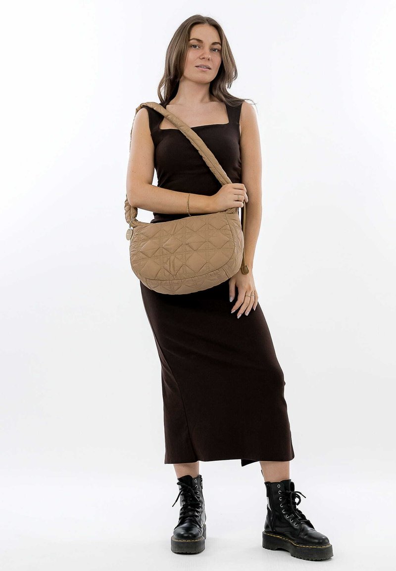 Quiltet beige skuldertaske med en buet form, flettet rem og subtilt diamantmønster. Modellen har en tætsiddende brun kjole og sorte støvler på.