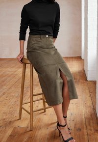 Femme assise sur un tabouret en bois, portant un haut noir à manches longues, une jupe midi olive avec fente à l'avant et des sandales à talons hauts noires.