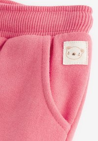 Vaaleanpunaiset puuvillaiset collegehousut, joissa on resorivyötärö, sivutaskut ja neliönmuotoinen etiketti karhukuvituksella. Sileä pinta ja rento malli.