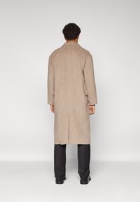 Beige Wollmantel mit geradem Schnitt, Reverskragen und Rückenschlitz, getragen über schwarzen Hosen und Schuhen. Rückansicht mit minimalem Detail.