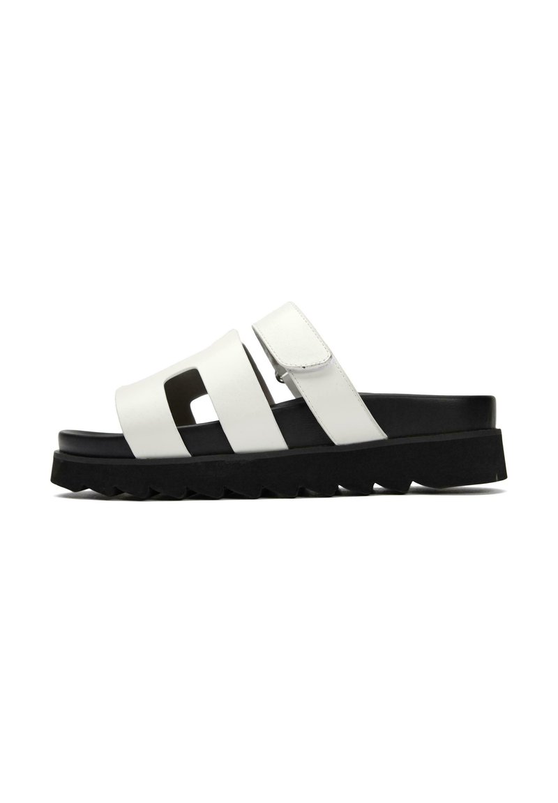 White sandal with two adjustable straps and cut-out design. Features a thick black sole with zigzag tread for grip and stability.