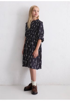 Dunkelblaues Kleid mit weißen Schleifenmustern, gerafftem Ausschnitt und Puffärmeln. Getragen mit braunen Schuhen und cremefarbenen Socken, vor einem hellen Hintergrund.