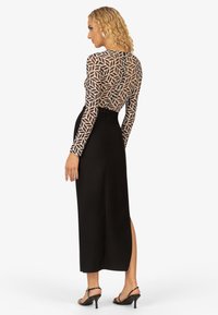 Langes schwarzes Kleid mit einem taillierten Oberteil und einem geometrisch gemusterten transparenten Überzug, mit langen Ärmeln und einem Schlitz an der Seite. Riemchen-Highheels in Schwarz.