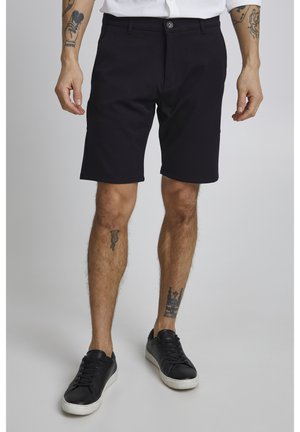 Homme portant un short noir sur mesure avec des baskets noires, debout devant un fond uni gris clair.