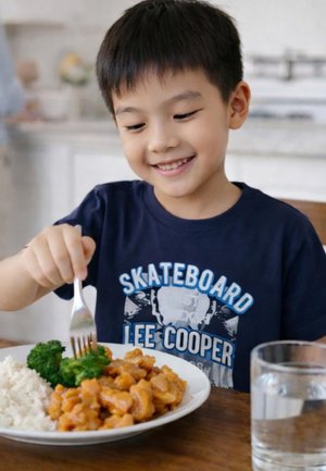 Jeune garçon souriant utilisant une fourchette pour manger du brocoli, avec une assiette de riz, poulet, brocoli et un verre d'eau sur la table.