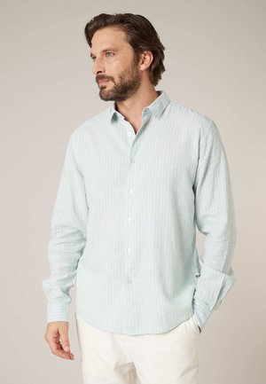 Homme barbu portant une chemise à manches longues à rayures bleu clair et un pantalon blanc, regardant vers la gauche sur un fond beige uni.