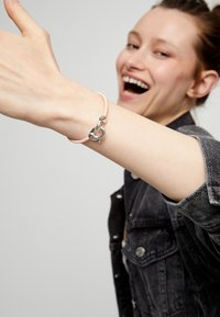 Charm en forma de corazón plateado en una pulsera rosa trenzada, llevada en un brazo de piel clara. El diseño texturizado contrasta con el charm liso.