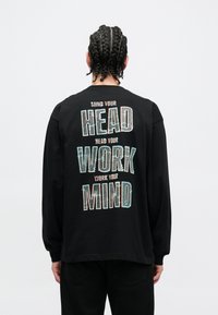 Schwarzes Langarmshirt mit großem, mehrfarbigem Text auf der Rückseite: "MIND YOUR HEAD, WORK YOUR MIND." Baumwollstoff, lockere Passform.
