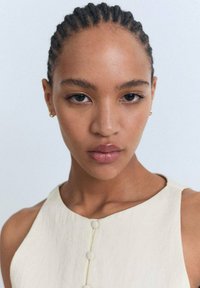 Nahaufnahme einer Person mit geflochtenem Haar, die ein cremefarbenes ärmelloses Oberteil mit Knopfakzenten trägt, vor einem glatten, neutralen Hintergrund.