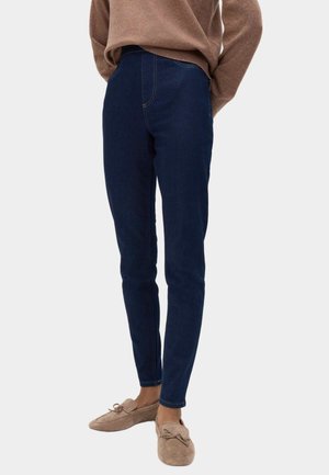 Personne portant un jean skinny bleu foncé, un pull marron clair et des mocassins en daim beige, debout devant un fond uni.