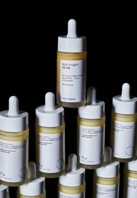 Gul glasflaska med pipett och vitt lock. Etiketten har svart text. Omgärdad av liknande flaskor mot en mörk bakgrund.