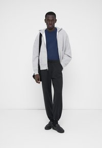 Sudadera con cremallera gris, camiseta azul, pantalones deportivos negros y zapatos negros sin cordones. Tejido suave con ajuste relajado y puños elásticos en los pantalones.