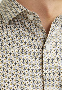 Camisa de botonadura frontal en tela blanca con un patrón geométrico en amarillo y azul marino. Presenta un cuello clásico y botones de plástico blanco.