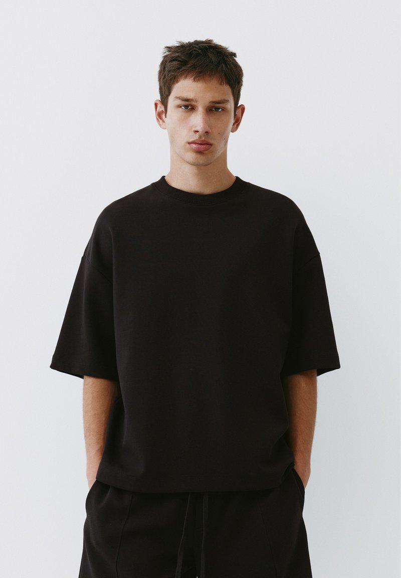 Jeune homme aux cheveux courts portant un T-shirt noir oversize et un pantalon noir, se tenant devant un fond clair uni.