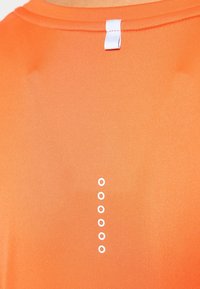 Camiseta naranja con cuello redondo, etiqueta de tela blanca en la parte trasera y cinco pequeños círculos blancos dispuestos en vertical.