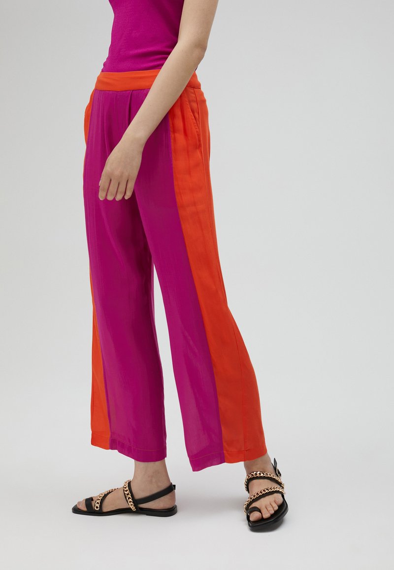 Vaaleanpunaiset ja oranssit leveälahkeiset housut, joissa on sileä pinta. Erityinen värikontrasti ja rento istuvuus, yhdistetty mustiin sandaaleihin.