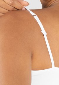 Weißes Tanktop mit einem Riemen, der mit einem Slider an einer glatten, braunen Hautschulter gesichert ist, wodurch die verstellbare Eigenschaft des Riemens betont wird.