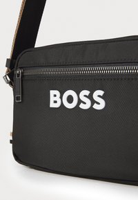 Čierna nylonová crossbody taška s uzatváraním na zips, štruktúrovaným povrchom, odvážnym bielym logom "BOSS" a kontrastným popruhom pre pohodlné nosenie.
