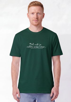 Homme aux cheveux rouge clair portant un t-shirt vert foncé avec un texte allemand blanc "Ich will ja nichts sagen, aber...", debout devant un fond uni.
