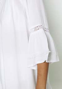 Blusa blanca con mangas acampanadas, que presenta un intrincado encaje y un ribete decorativo en el dobladillo. Fabricada con tejido ligero.