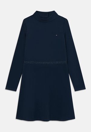 Robe bleu marine à manches longues avec un col haut, présentant un corsage ajusté, une jupe évasée et un discret logo près de la poitrine. Texture de tissu douce.