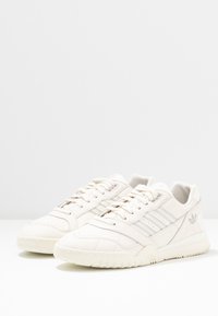 Baskets en cuir de couleur crème clair avec une finition texturée, présentant des accents argentés et un design classique à trois bandes sur les côtés.