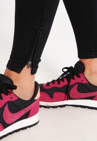 Jeans negros con un detalle de cremallera en el tobillo, combinados con zapatillas Nike negras y fucsias que presentan texturas de malla y gamuza, cordones negros y suelas de goma.