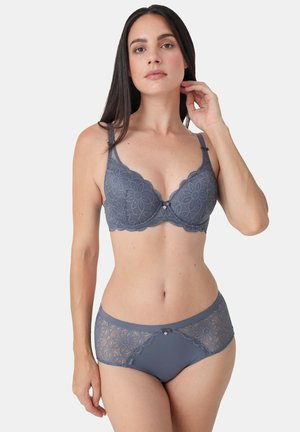 Woman with long dark hair wears matching gray floral lace bra and hipster panties, posing against a plain light background.