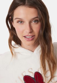 Hvid hoodie med et blomsterbroderi i rød og marineblå, med gyldne bogstaver. Blød tekstur, med hætte og afslappet pasform.