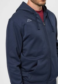 Tumesinine kapuutsiga tõmblukuga jakker, mis on valmistatud puuvillasegust, omab paelu reguleeritavas kapuutsis, eestaskuid ning väikest valget logo vasakul rinnal.