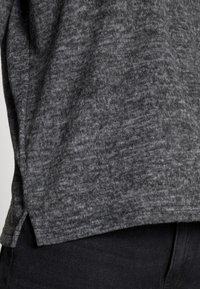 Tessuto a maglia grigio scuro con una superficie texture, che mostra una cucitura laterale e una leggera spaccatura all'orlo. Abbinato a pantaloni neri.